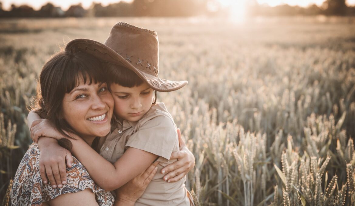 Smiling mom embracing cute son wearing hat while sitting in agricultural field in sunny evening at sundown