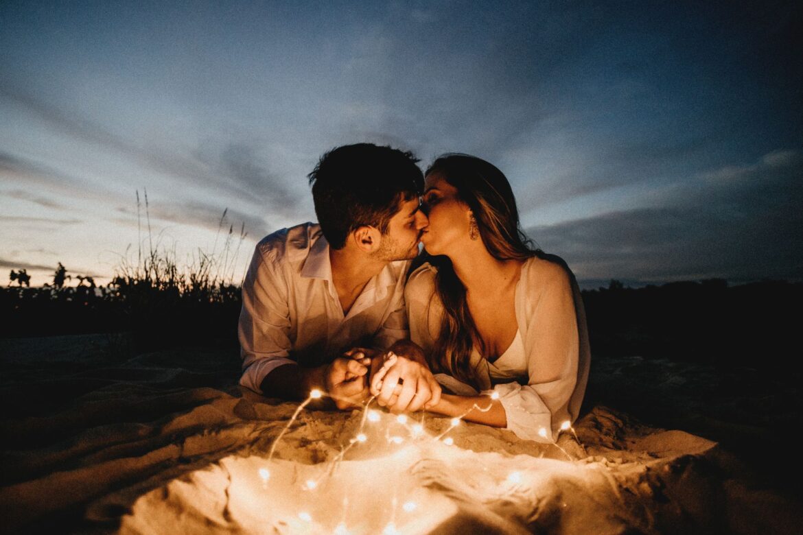 Couple kissing and holding hands while lying near illuminating garland against picturesque sky