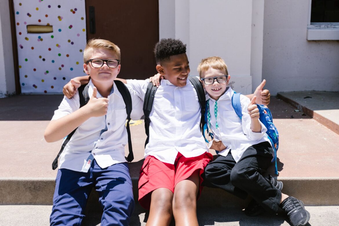Three smiling boys with backpacks sitting on school steps, showing thumbs up.
