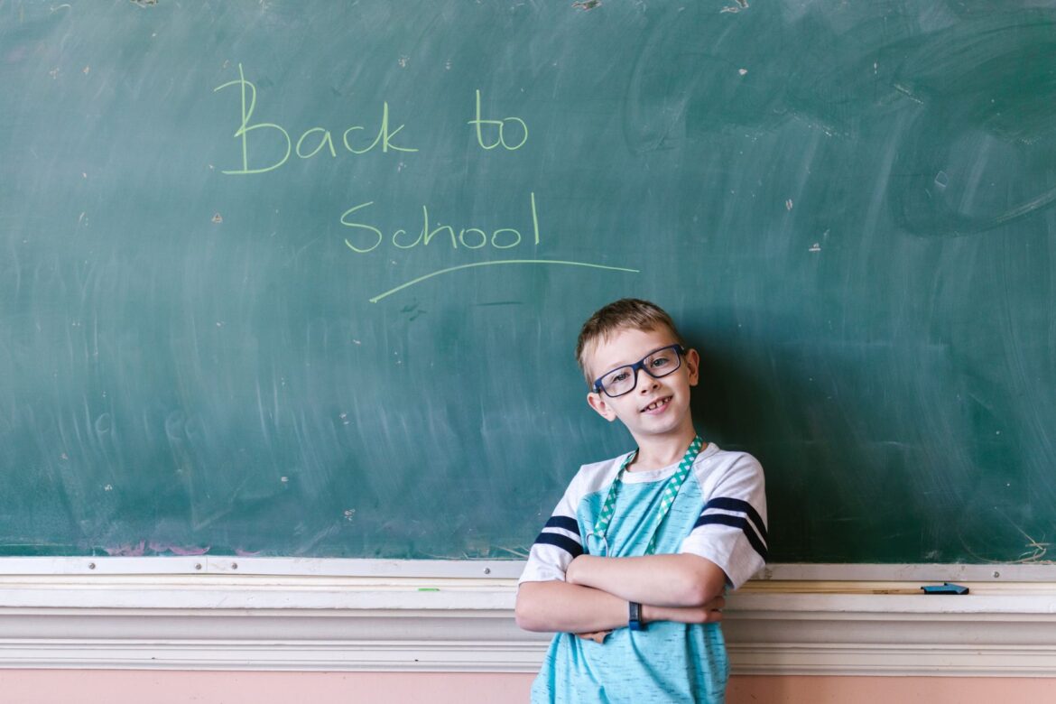 A young boy stands confidently with folded arms in front of a chalkboard saying 'Back to School'.