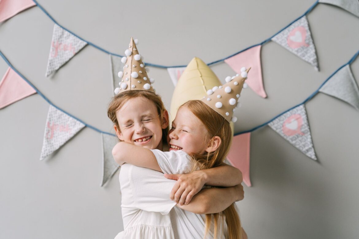 Two girls hugging at a lively indoor birthday party with festive hats and banners.