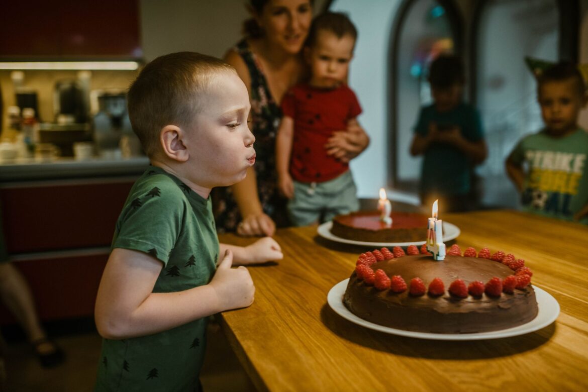 Young boy celebrating birthday by blowing candles on chocolate cake with raspberries.