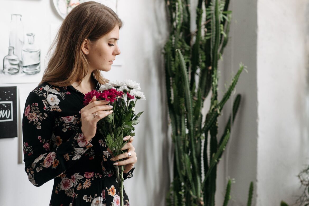 A young woman in a floral dress holding flowers indoors, standing near a cactus plant.