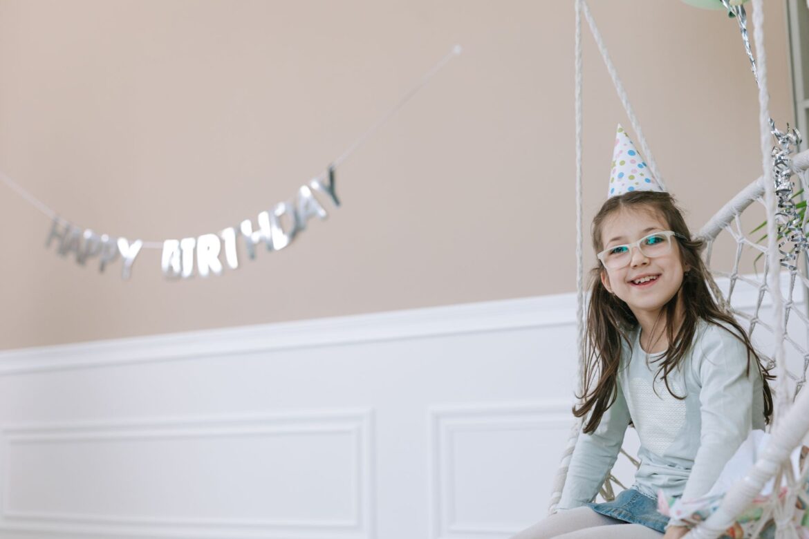 Young girl wearing glasses and party hat smiling on a swing indoors.