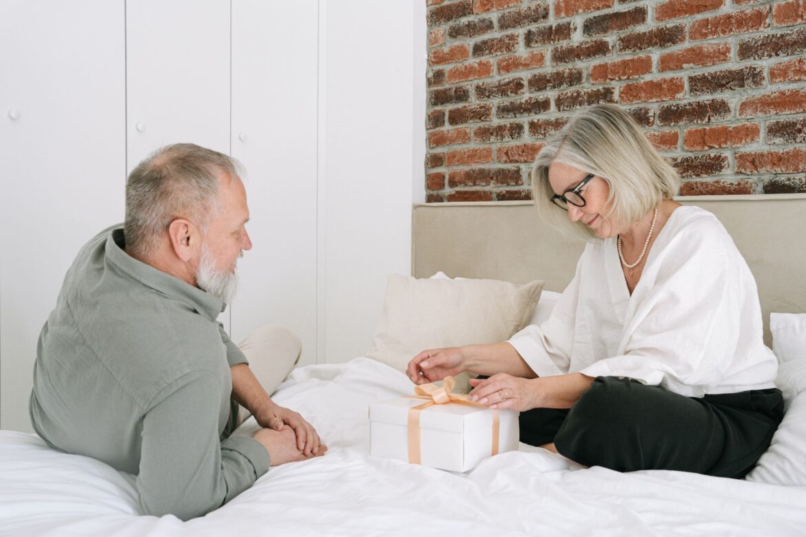 Senior couple sitting on bed exchanging a wrapped gift, showcasing love and companionship.